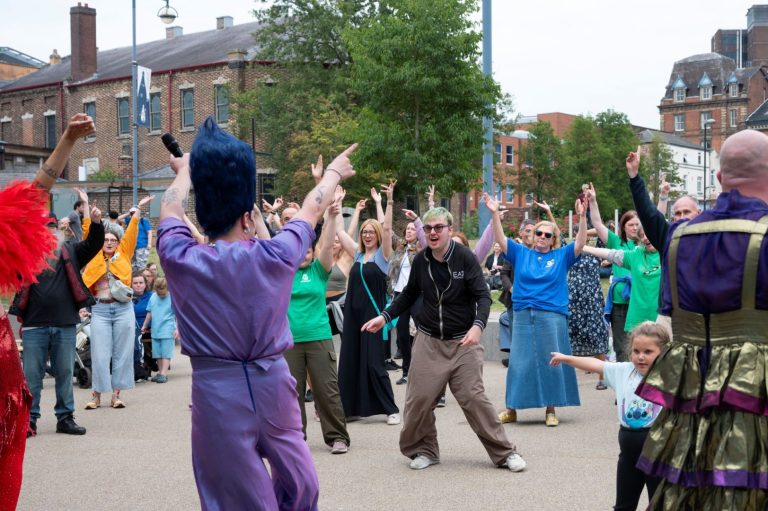 The image is taken outside on a street with buildings in the background. The backs of three performers are at the front of the image. The central performer is wearing a purple top and trousers and large purple wig. The performers are looking over to lots of people who have their arms in the air and are dancing.