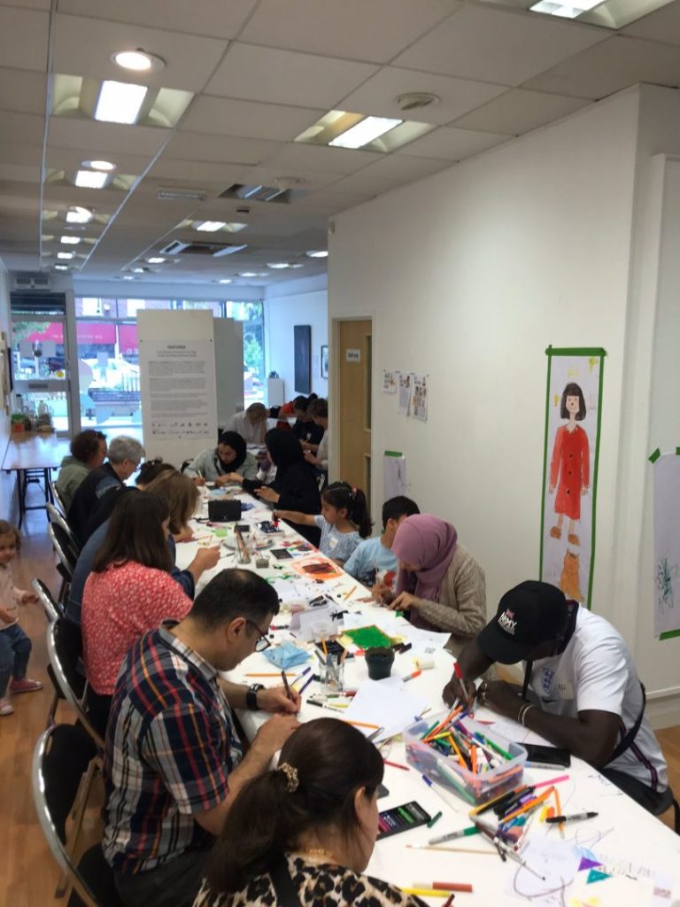 The image shows a central large table with lots of paper and materials on. A number of people are sat and standing around the table working on various activities.