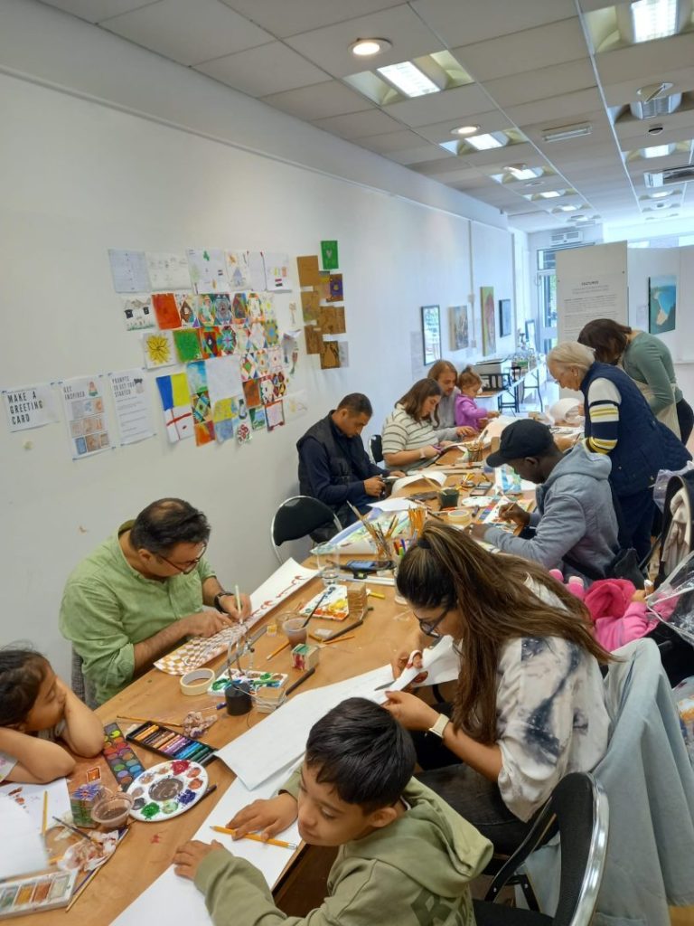 The image shows a central large table with lots of paper and materials on. A number of people are sat and standing around the table working on various activities.
