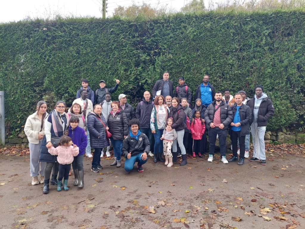 A large group of people are standing to have their photograph taken outside. There is a large hedge in the background. The group includes children and adults.