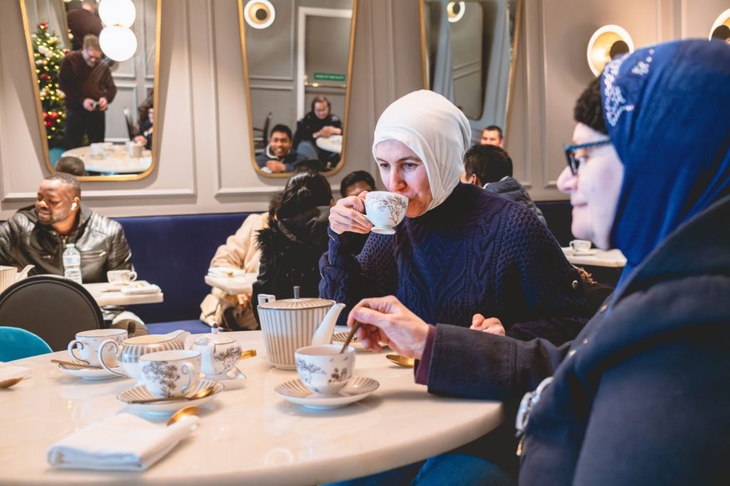 The photograph is taken inside and shows two people sat at a table, with other people in the background. The table is set with cups, saucers and tea pots and one of the people is taking a sip from a tea cup.