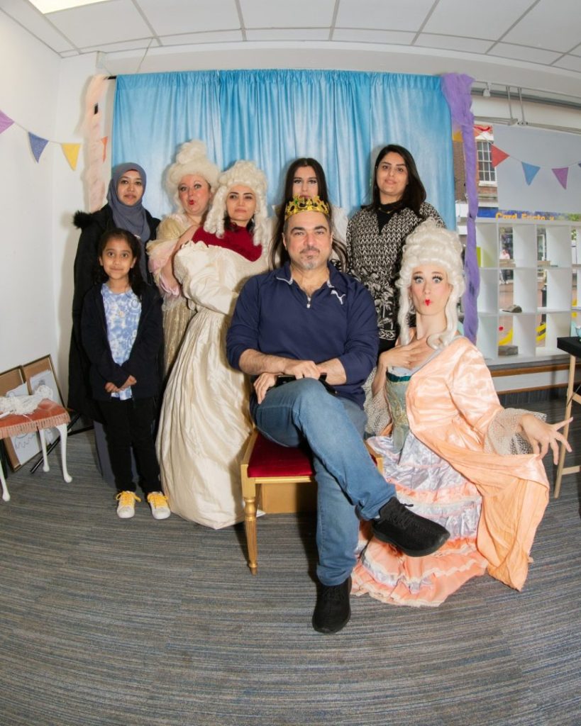 Eight people are posing to have their photograph taken inside a room against a blue backdrop. Three of the people are wearing costumes of long dresses and white wigs. The central figure is seated with the other people arranged around them.