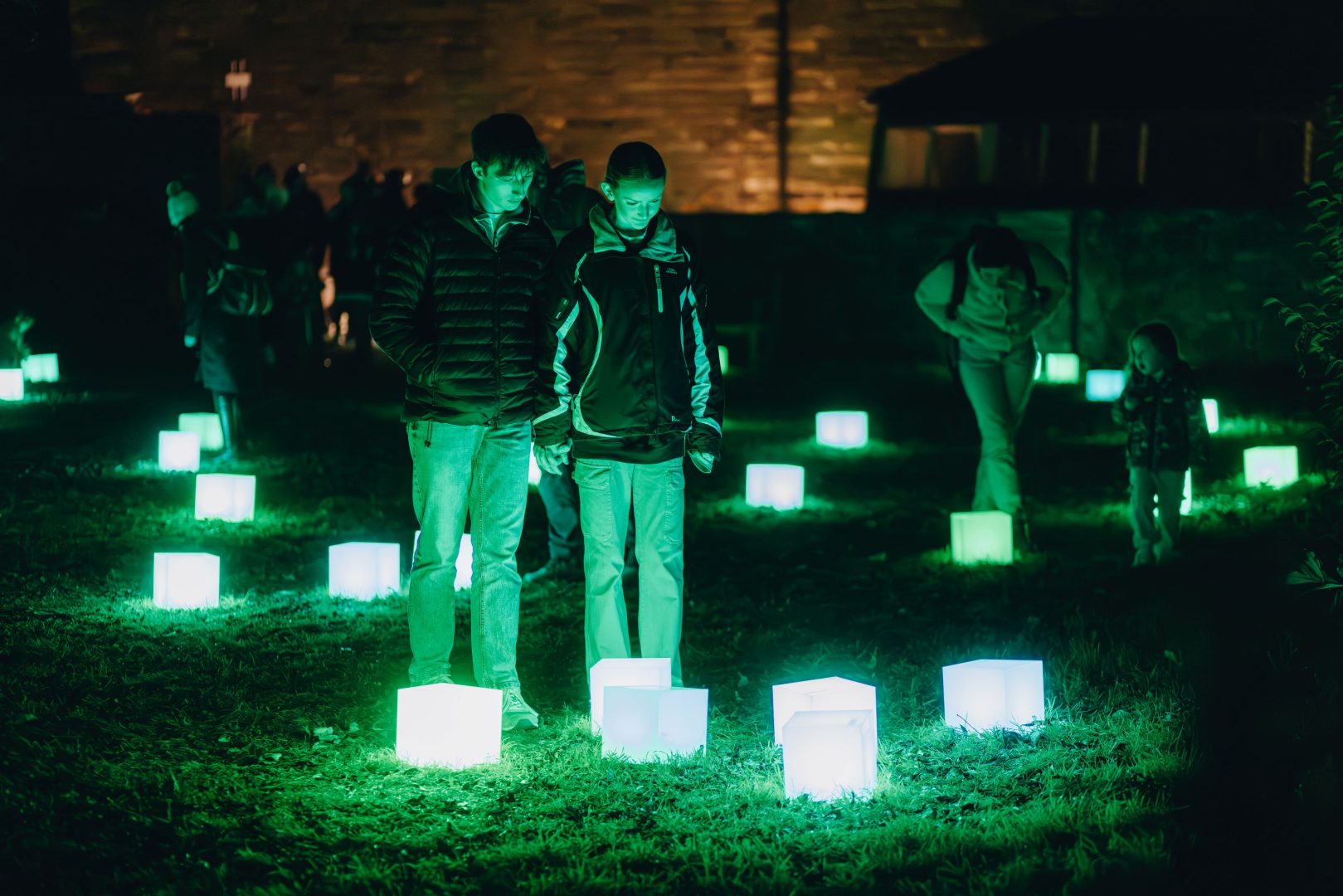 Two young adults are standing on the grass in the dark looking down at several small boxes on the ground that are glowing bright green. There are more boxes dotted around the floor in the background and several more people.