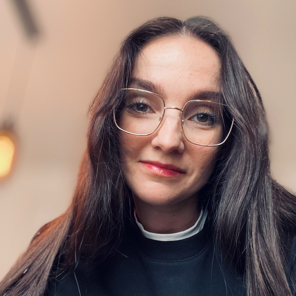 A white woman with long, dark, centre-parted hair, large, round, metal-framed glasses, and a warm smile, looks directly at us.