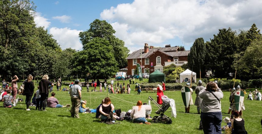 Brampton museum is shown in the distance with a large grassed area in the foreground. People are on the grass and there are some gazebos. Two knights have their back turned revealing brightly coloured shields.