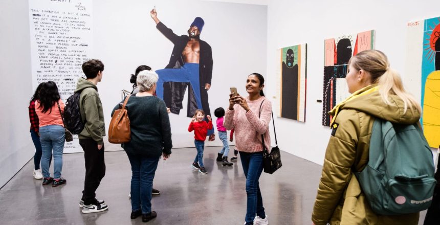 A group of people are looking at images inside of a gallery. There are three colourful paintings on the right hand wall. On the back wall, there is a large image of a person.