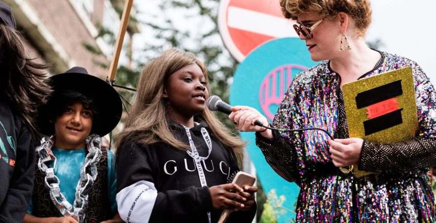 Comedian Mavis Davis is interviewing a you g person. Mavis has a sequined multi-coloured dress, yellow clipboard, Large glasses and curly hair piled on top of her head. She is holding a hand-held mic towards a girl who is wearing a long caramel blonde wig, and wearing a black Gucci hoodie, holding a long-handled hair brush. To her side a boy has also clearly hit the dressing up box and wears a black wide-brimmed trilby, blue tie dye T and an exaggeratedly large silver chain that looks to be made from foil-covered cardboard.
