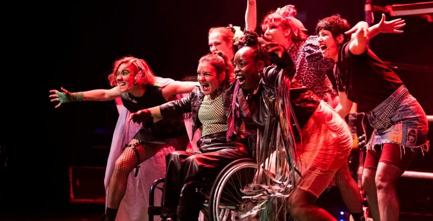 The image is taken inside with a dark background and looks like it is on a stage. It looks like the stage is lit with red lights. The image captures a group of six performers, three at the front, three at the back, all with outstretched arms and looking like they are singing. They are all looking towards the left, out of shot where the audience must be. The performers are wearing different brightly coloured clothing and leather jackets, dresses or tops. The central performer is using a wheelchair and is wearing black leather trousers, a black jacket and a green top.
