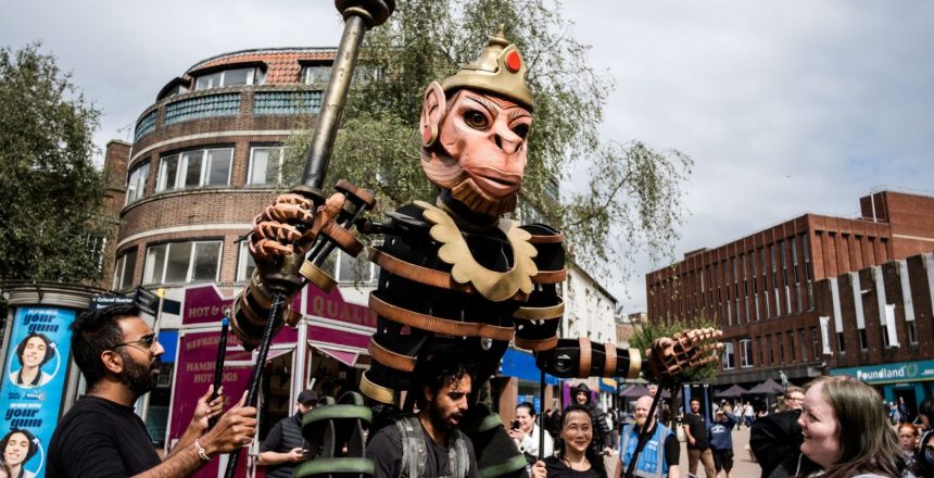One of the characters from Ancient Giants, can be seen interacting with an audience member, who is looking at it and smiling. The character towers above them. The scene is taken on a street with buildings in the background.