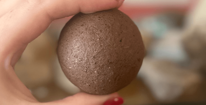 The image shows a Dorodango ball held between a person's thumb and index finger. The ball is made from soil, so is brown in colour and has a soil-like texture.
