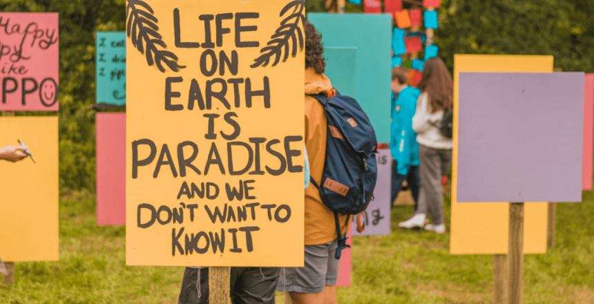 Multicoloured placards a placed in a field. Their is a person with a backpack stood among them. The placard at the very front is orange with black writing that reads 'Life on Earth is paradise and we don't want to know it'