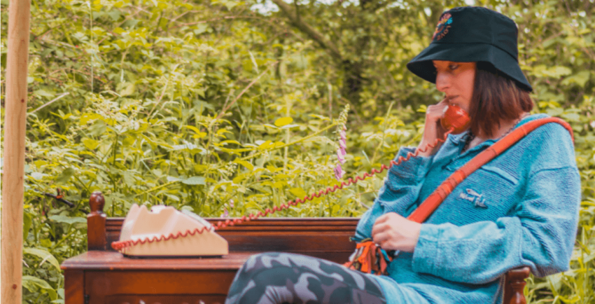 A person wearing a bucket hat and blue jumper is sat onsite on a wooden bench. They are holding the receiver of an old fashioned telephone.