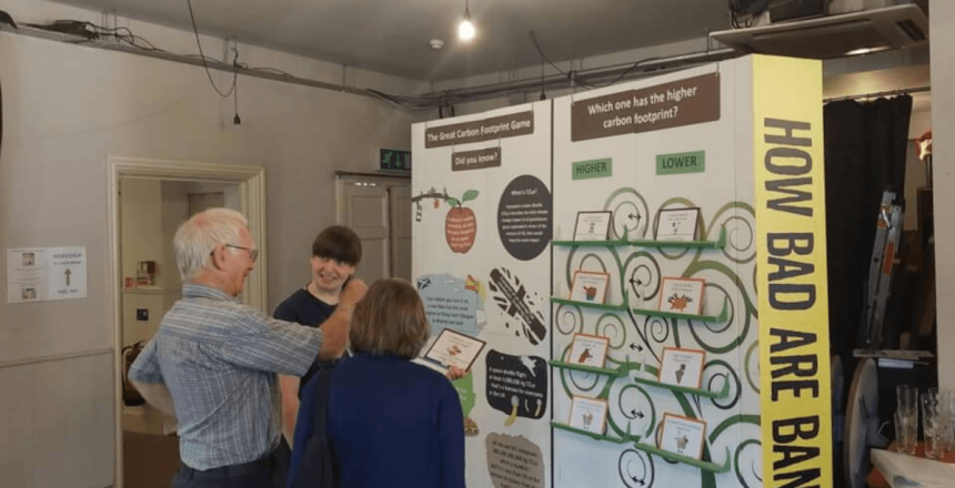 Three people are stood indoors beside a large board which has words and illustrations on it. On the side on a yellow background with black writing reads 'How Bad Are Bananas?'