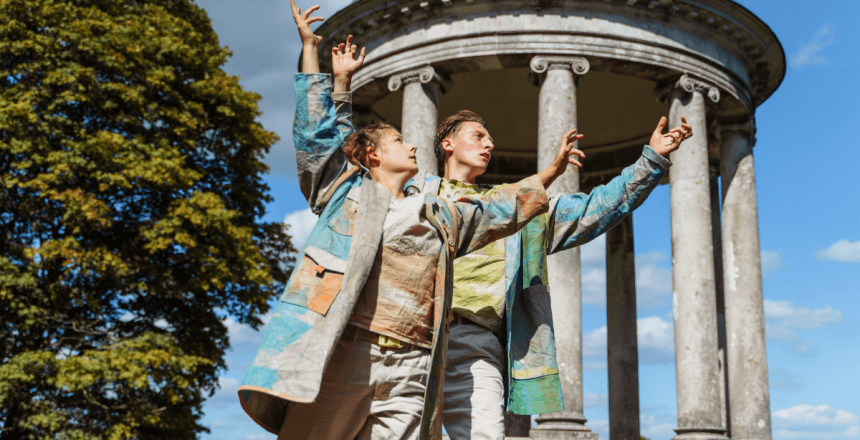 Two people are stood outside. They are both holding their arms up, one behind the other. Behind them is a concrete statue with columns. There is also green trees and the sky is blue.
