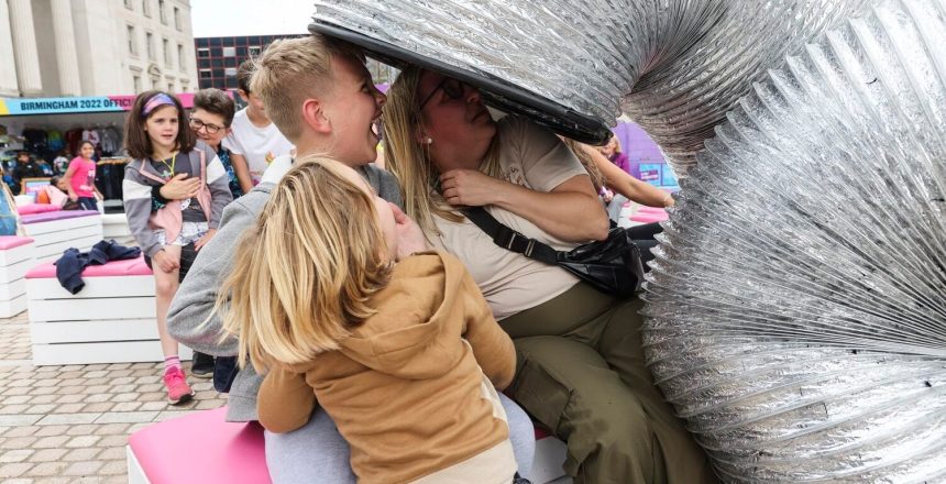 The image shows a human sized silver slinky, towering over three people as though they might go inside. The people are laughing.