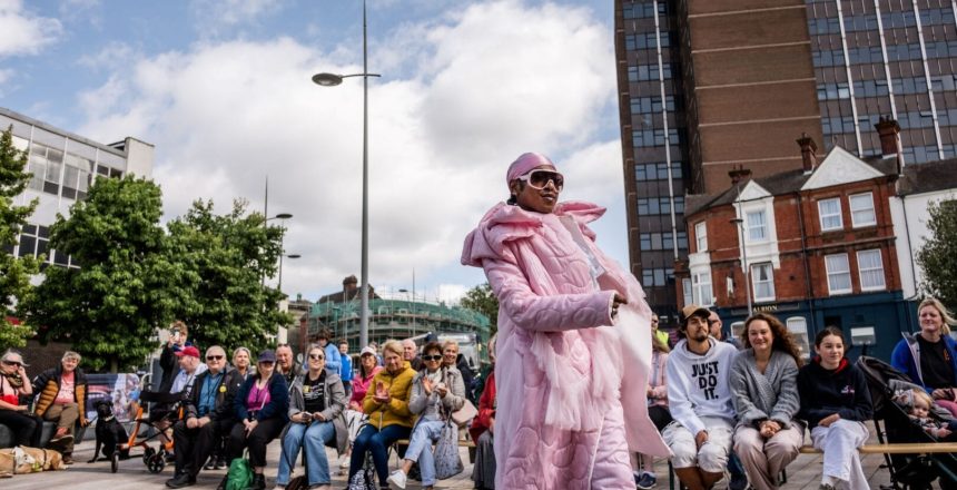 A performer dressed in pink trousers, scarf and coat and wearing sunglasses, is walking towards the right of the photo. Rows of people can be seen in the background sitting down and watching the performer walk past. The photo is taken in a city centre outside, with buildings in the background. It is a sunny day.