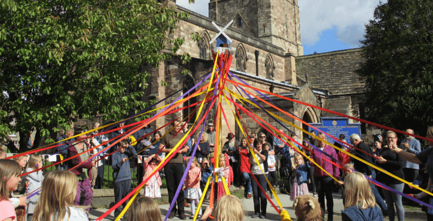 A large audience of people are gathered outside of a church. They are gathered around a maypole, with some holding a piece of brightly coloured ribbon attached to the pole.