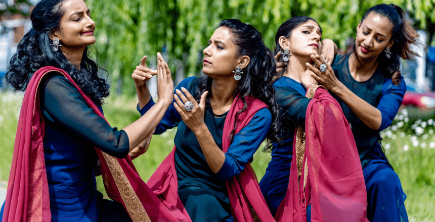 There are four performers stood outdoors. There are green trees and grass in the background. They are performing in pairs, gesturing towards one another. They are wearing royal blue dresses and are draped in a burgundy coloured scarf with gold edging.