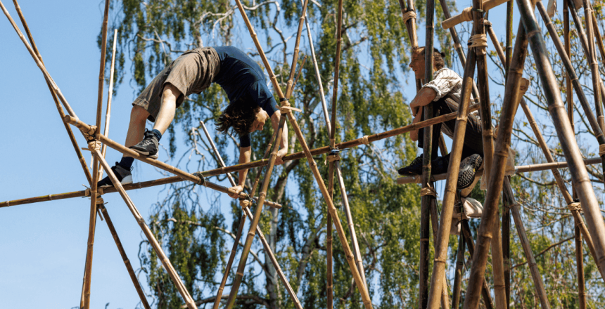 A person is in a crab position, almost like a backflip, within some strong pieces of bamboo. There is a person crouched within the bamboo next to them.