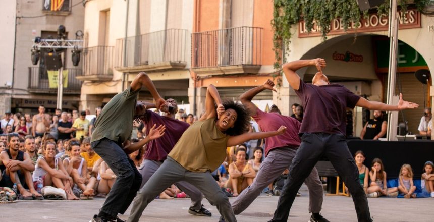 A group of five black dancers are moving energetically in an outdoor space. A woman is standing with her legs wide apart, punching out to the side with her left arm. Her right arm is bent inwards and her hand her is making a fist with a thumbs down sign. Her expression is wide eyed and she looks like she is screaming. Around her four men are bending over backwards with similar poses. Behind them a large crowd of spectators are sitting on the floor or standing. They are all wearing dark trousers and trainers, and t-shirts in muted tones.