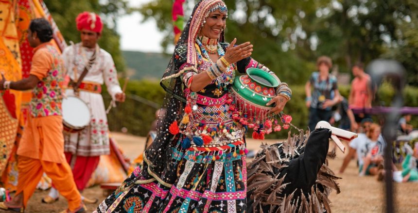 A smiling lady is in the open and looks to be mid-performance, engaging with people off-camera. She is in a stunningly vibrant costume of floor-length dress and long, flowing headscarf. The base colour of these pieces is black but they are highly adorned with trim, beads, tassels and stitching in pink, turquoise, orange and white. She has a beaded choker at her neck and both arms are adorned with lots of bangles. She is holding a large green pot under her left arm, while her right arm is extended to the unseen crowd. To her right is a large, naive puppet of a bird made from black fabric with brown feathers attached and a wooden beak. The background is blurred but you can see a few people sitting and standing in the audience, plus the edge of an orange, red, and yellow fabric tent with two men in colourful Indian dress outside.