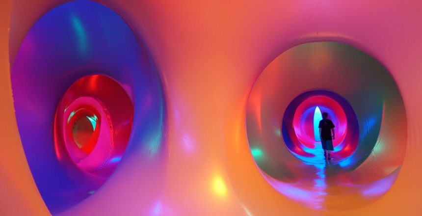 We are looking at the interior of a Luminarium, an inflatable structure with corridors and chambers bathed in coloured light. From this view we are standing in the intersection of two corridors that can both be seen down. The corridors are circular and oval in shape and undulate side to side as they progress. The foreground where we are is a soft peach colour, and you can see the colours morph and blend as your eye travels down the corridors into blues, pinks and reds with purple and green spotlights at points adding pattern. You can see the black silhouette of a lone figure walking down the end of corridor on the right.