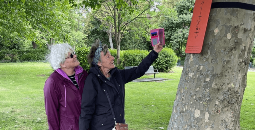 Two people are outdoors. One is holding their phone up to a red plaque attached to a tree.