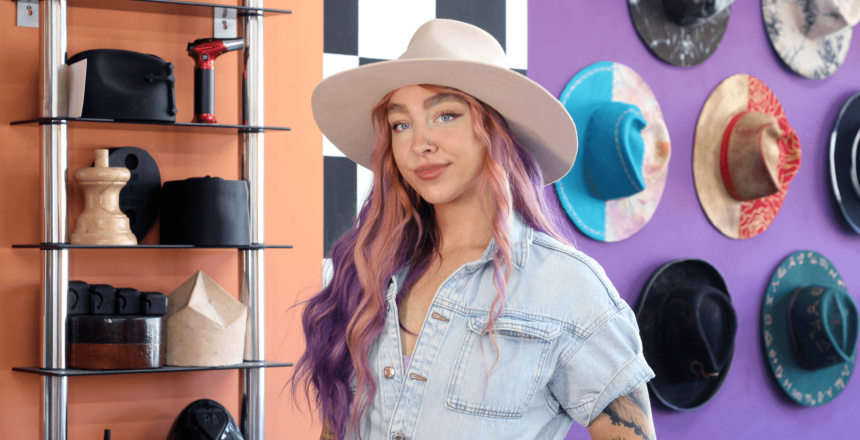 Holly is looking into the camera. She has pink and purple hair and is wearing a white hat. Behind her are purple and orange walls, and different, colourful hats are hung onto the wall.