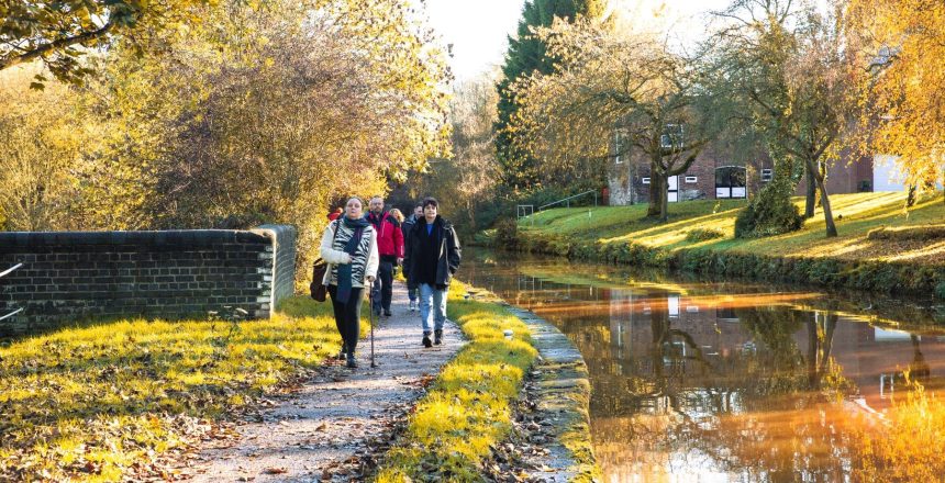 The group of walkers are coming towards us in a line along the towpath. To the right is the canal and on the opposite bank are trees, a grass slop and a building in the background. The trees and building are reflected in the sunshine onto the orange water.