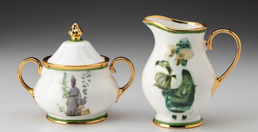 A ceramic sugar bowl and milk jug are set against a soft grey background. The items are mainly white with gold rims at the top and bottom, handles, and a thin line of emerald-green paint next to the gold. The pointed finial on the lid on the sugar bowl is gold with a ring of green at its base. There is printed imagery on each piece in muted tones. On the bowl, a lady in a purple empire-line dress and matching hat is surrounded by shoots and tendrils of greenery and pink flowers. The jug has a figure in a long white dress with a blurred image of people, including one riding a horse. The figure is holding large leaves across their right shoulder, partly obscuring their face.