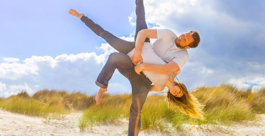 Two acrobats dressed in blue jeans and white t-shirts are performing on a sandy beach under cloudy blue skies. They are in motion as the man twirls the woman around so that her head is close to the floor and her legs are raised against the sky in a V shape. The woman’s long, blonde hair is wheeling out behind her.