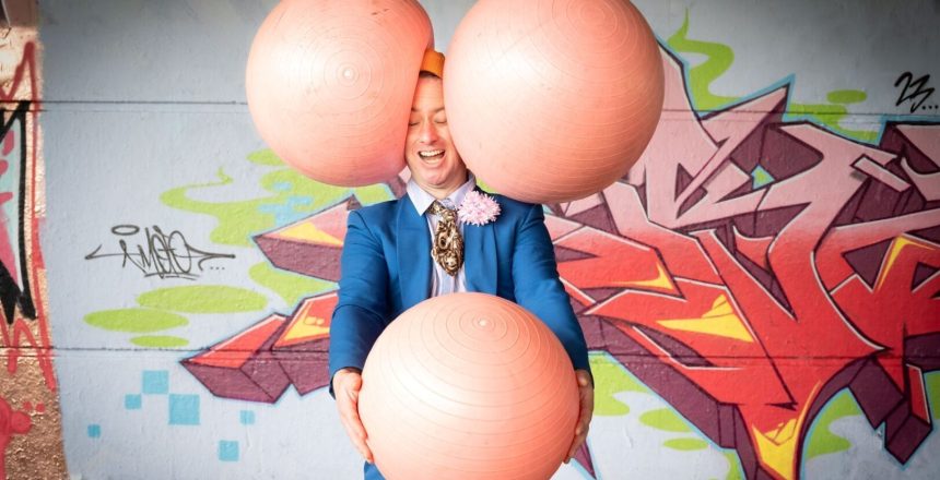 We are seeing a man at the exact moment that his head is being squished between two large, pink rubber balls that have been thrown at him. His eyes are closed, and his mouth is open in mid-laugh. He holds a third ball in his hands stretched in front of him. He’s wearing a blue jacket, light pink trousers and a very short brown patterned tie with a pink flower in his buttonhole. He is standing in front of a white wall with large, highly stylised graffiti tags.