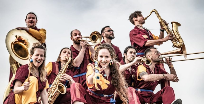 Nine people dressed in red-and-gold costume are seen in close-up grouped together on a pebble beach. The front row are sitting and the other standing behind. Some hold brass instruments of all sizes and some are blowing into them. They look happy. One woman centre front is holding a staff wrapped in red and gold cloth directly outstretched to the viewer.