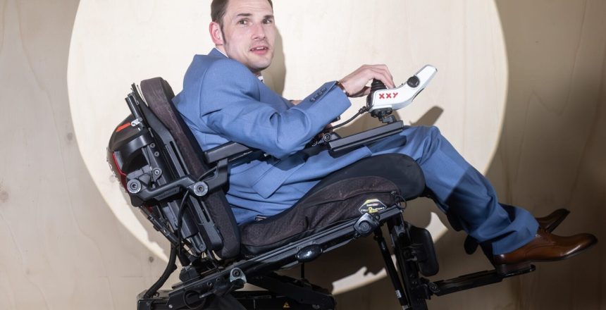 Shaun is in right-side profile. He is a white man with dark hair wearing a petrol-blue suit. He is spotligt against the backdrop of a wall in a neutral colour. Shaun is sitting in his wheelchair.
